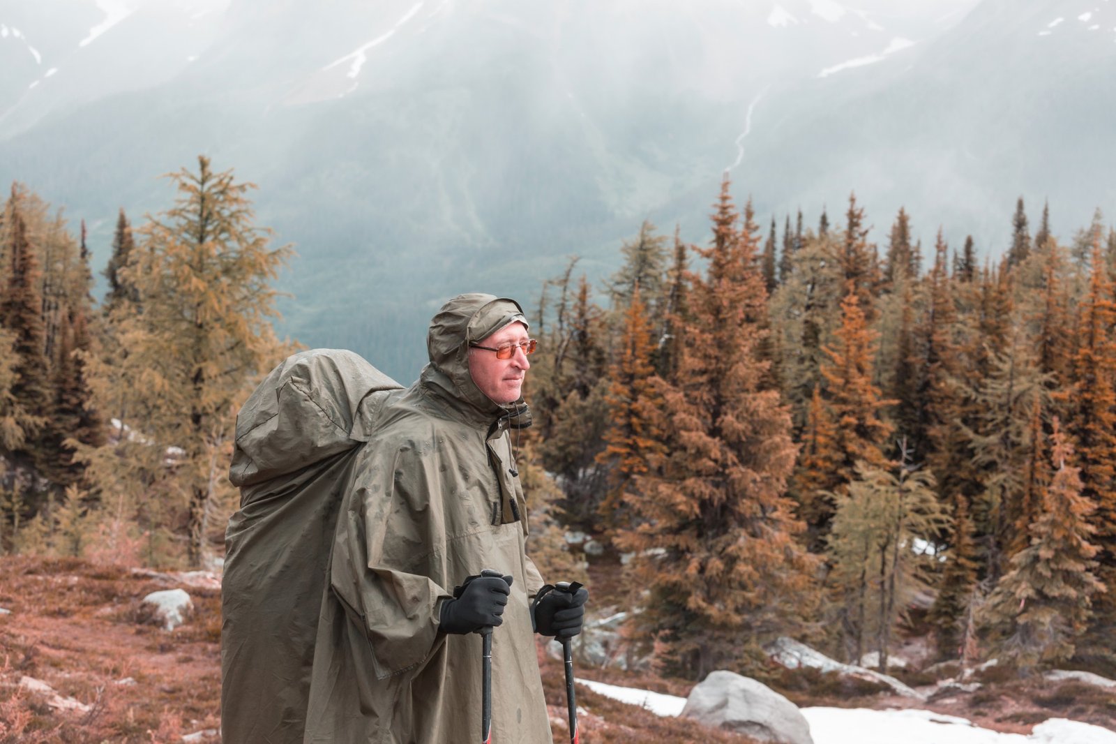 Randonneur en poncho kaki qui couvre aussi son sac à dos, devant une forêt de mélèzes